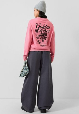 Vrouw met grijze muts, roze sweatshirt met zwarte bloemmotief tekst op de achterkant, brede donkere broek en houdt een kleine blauwe handtas vast.