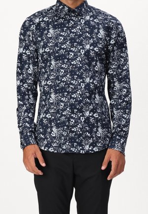Camisa azul marino de manga larga con botones y estampado blanco de flores y pájaros, usada con pantalones negros.