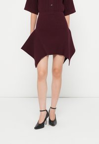 Burgundi színű, bordázott kötésű ruha rövid ujjakkal, aszimmetrikus szegéllyel, szövettextúrával, fekete bokapántos magassarkú cipővel párosítva.