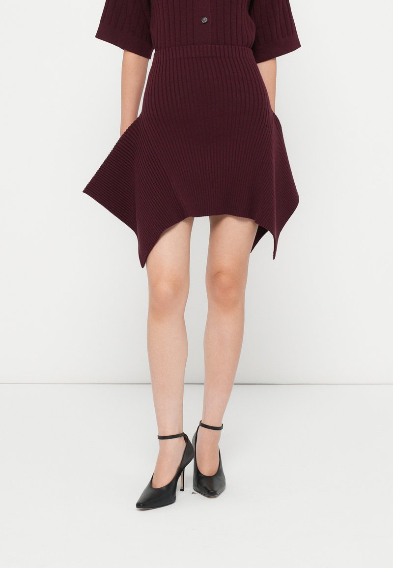 Burgundi színű, bordázott kötésű ruha rövid ujjakkal, aszimmetrikus szegéllyel, szövettextúrával, fekete bokapántos magassarkú cipővel párosítva.