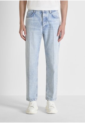 Uomo con jeans azzurri a gamba dritta e sneakers bianche, in piedi contro uno sfondo chiaro e uniforme.