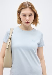 Νεαρή γυναίκα με κοντά κόκκινα μαλλιά φοράει ένα ανοιχτό μπλε t-shirt BOSS και κρατάει μια μπεζ τσάντα στον ώμο της, σε απλό φόντο.