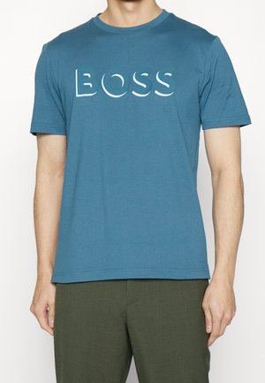 Mann, der ein blaues T-Shirt mit dem Wort "BOSS" in großen weißen Buchstaben auf der Brust und dunkelgrüne Hosen trägt.