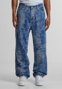 Blue denim jeans with a loose fit, featuring a textured pattern and contrasting stitching. Styled with white sneakers.