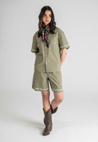 Olivenfarget kortermet skjorte og shorts-sett, med blomsterbroderi, kombinert med et mønstrete halsbånd og brune cowboy-støvler.