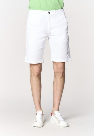 Uomo con pantaloncini bianchi fino al ginocchio, sneakers bianche con dettagli rossi e una camicia verde chiaro, in piedi davanti a uno sfondo neutro.