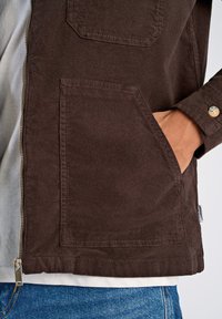 Chaqueta de pana marrón con cremallera, dos bolsillos frontales y un corte relajado, superpuesta sobre una camisa de color claro y jeans azules.