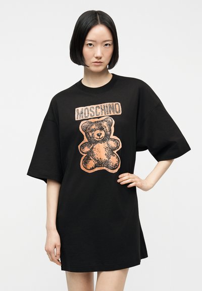 Νεαρή γυναίκα με κοντά μαύρα μαλλιά που φοράει ένα υπερμεγέθες μαύρο φόρεμα T-shirt Moschino με γραφικό αρκουδάκι.