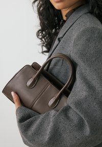 Borsa a tracolla in pelle marrone con due manici arrotondati, caratterizzata da una forma rettangolare e cuciture minime, tenuta contro una manica grigia testurizzata.