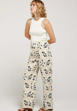 Femme aux cheveux blonds portant un haut blanc sans manches et un pantalon taille haute à larges jambes avec des motifs floraux noirs et géométriques beige.
