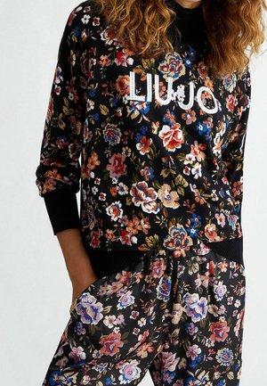Kobieta ubrana w czarną, kwiatową bluzę i spodnie z kolorowymi kwiatami oraz napisem "LIU JO" na bluzie.