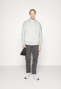 Hanorac gri cu buzunar kangaroo, asortat cu pantaloni cargo negri și adidași albi. Modelul ține o geantă mică neagră; texturi de țesătură netedă.