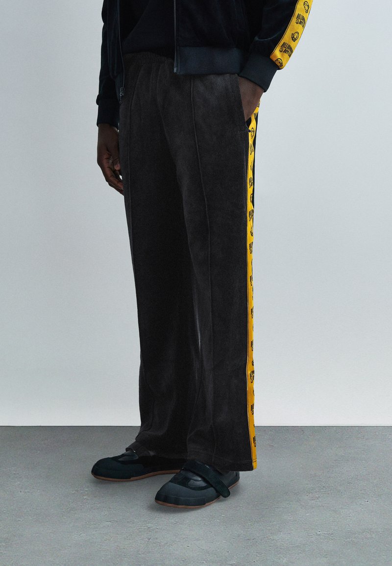 Μαύρο βελουτέ παντελόνι φόρμας με  κίτρινες πλευρικές ρίγες που φέρουν γραφικό σχέδιο, συνδυασμένο με μαύρα παπούτσια τύπου slip-on. Ελαστική μέση, άνετη γραμμή.