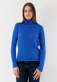 Maglione blu a collo alto realizzato in morbido tessuto a maglia, con maniche lunghe e vestibilità rilassata. Indossato con jeans blu scuro su uno sfondo chiaro.