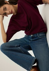 Burgundowy luźny top w połączeniu z szerokimi niebieskimi dżinsami. Tkanina wydaje się być miękka, z gładką fakturą i subtelnymi detalami szycia.