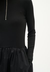 Rochie neagră cu mâneci lungi plisate și fermoar la gât, având un top ajustat și o fustă voluminousă, adunată.