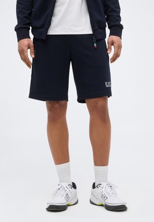 Muž nosiaci čierne šortky EA7 Emporio Armani, biele ponožky do členkov, biele tenisky EA7 a námornícku bundu s bielym tričkom pod ňou.