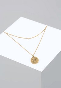 Deux colliers en or superposés sur un cube blanc, l'un avec de petits perles et l'autre avec un pendentif rond gravé.