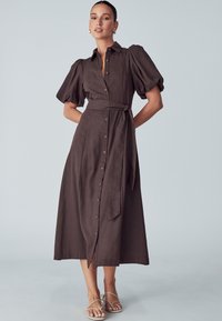 Robe midi marron à boutons avec manches bouffantes, taille cintrée et détail ceinturé. Texture lisse et silhouette droite. Assortie à des sandales à lanières.