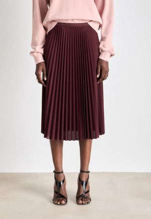 Vrouw draagt een bordeauxrode geplooide midirok, lichtroze top met lange mouwen en zwarte sandalen met hoge hakken en bandjes, staand op een beige vloer.