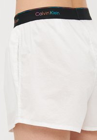 Vita bomull boxershorts med svart elastiskt midjeband med "Calvin Klein" i flerfärgad text. Slät textur, avslappnad design.
