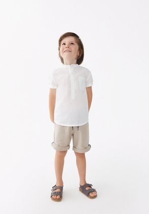Camisa de lino blanca de manga corta con un bolsillo, pantalones cortos beige enrollados y sandalias grises con hebillas. Un atuendo simple, ligero y transpirable.