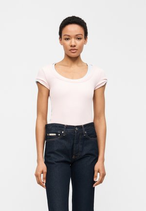 Femme aux cheveux courts et bouclés portant un haut à manches courtes rose clair rentré dans un jean taille haute bleu foncé.