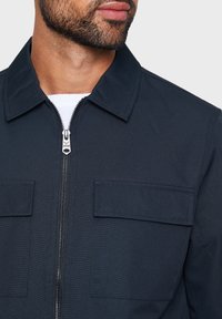 Mørk marineblå jakke lavet af glat stof. Har krave, lynlås foran og to brystlommer. Enkel design uden synlige mønstre.