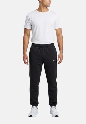 Uomo che indossa una maglietta bianca semplice, pantaloni della tuta neri con vita elastica con logo Champion e scarpe da ginnastica bianche, in piedi contro uno sfondo semplice.