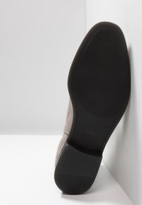 Semelle en caoutchouc noir d'une chaussure en daim marron clair, inclinée contre un mur blanc, montrant la texture de la semelle, le logo Hugo et la taille 41.