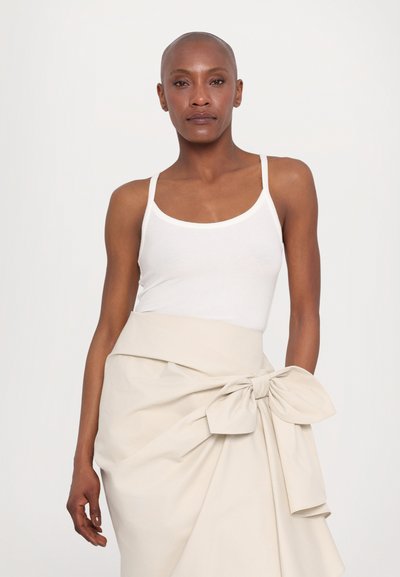 Débardeur blanc à fines bretelles associé à une jupe beige clair ornée d'un grand nœud à la taille, en tissu lisse et au design fluide.