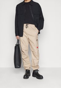 Beiga cargobyxor med dragsko i midjan, svart skjortjacka och svarta läderstövlar. Modellen håller en svart tote-väska.