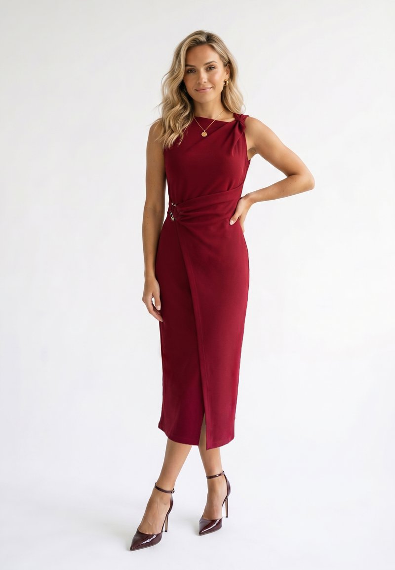 Femme aux cheveux blonds ondulés portant une robe bordeaux sans manches au genou et des talons assortis, debout avec une main sur la hanche.