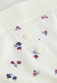 Zbliżenie na biały materiał z drobnymi czerwonymi i ciemnoniebieskimi kwiatowymi wzorami oraz dwoma białymi guzikami na przeszytym plisku.