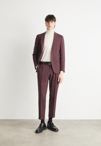 Burgundy suit with tailored fit, paired with a beige turtleneck. Black belt and shoes complete the look. Smooth fabric with minimal detail.