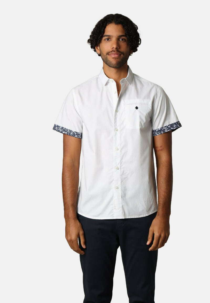 Chemise blanche à manches courtes avec une bordure bleue à motifs sur les manches et une poche poitrine. Dotée de boutons sur le devant et d'un petit détail de logo.