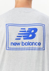 Grå bomull t-skjorte med blå grafisk design som har tekst med "new balance" og et stilisert "N"-logo innenfor en rektangulær ramme.