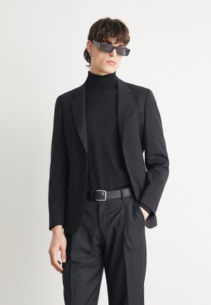 Blazer sartorial nero sopra un dolcevita nero, fit regolato con spalle strutturate, abbinato a pantaloni neri e una cintura nera.