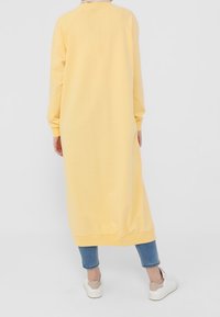 Vestido amarillo de manga larga hecho de una tela suave, con un ajuste holgado y puños acanalados, combinado con jeans azules y zapatillas blancas.