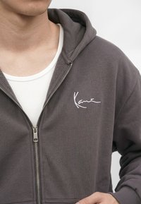 Persoană purtând un hanorac gri închis cu fermoar parțial și logo alb "Kani" și un tricou alb cu dungi sub el.