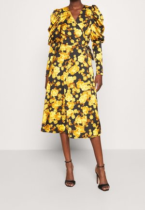 Robe portefeuille noire à imprimé floral jaune, manches longues bouffantes, longueur midi, portée avec des escarpins noirs à bride cheville sur un fond clair.