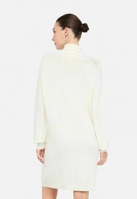 Abito in maglia crema a maniche lunghe, lungo fino al ginocchio, con colletto alto e delicati dettagli testurizzati, completato da un piccolo bottone sulla parte posteriore.