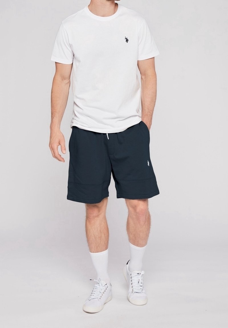 Biała bawełniana koszulka z małym czarnym logo, w połączeniu z granatowymi szortami z miękkiego materiału, z troczkiem, noszona z białymi skarpetkami i trampkami.