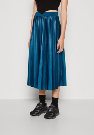 Kvinde iført en tealblå plisseret midinederdel, sort cropped top, sorte strømper og sorte atletiske sneakers, stående på en grå gulv.