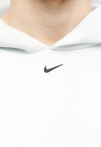 Close-up van een persoon die een witte hoodie draagt met een zwart Nike-logo gecentreerd onder de halslijn.