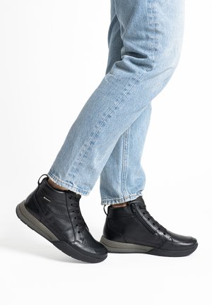 Füße mit schwarzen Leder-Stiefeletten mit seitlichen Reißverschlüssen und Schnürungen, hellblaue Jeans leicht hochgekrempelt vor weißem Hintergrund.