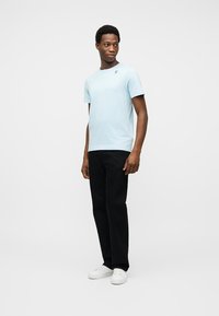 Άνδρας που στέκεται, φοράει ανοιξιάτικο μπλε κοντομάνικο πουκάμισο, μαύρο παντελόνι και λευκά αθλητικά παπούτσια, κοιτώντας ελαφρώς προς το πλάι.