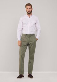 Camicia button-up a righe rosa chiaro con maniche lunghe, abbinata a pantaloni chino verde oliva e scarpe slip-on marroni scure. Tessuto morbido, vestibilità su misura.