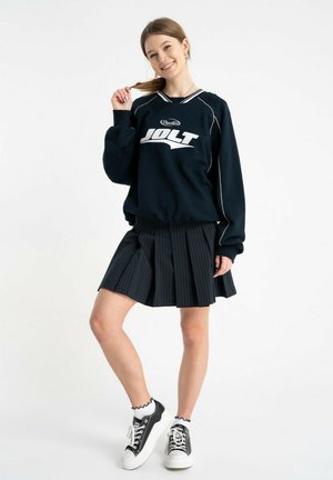 Uśmiechnięta kobieta w czarnej bluzie "JOLT", plisowanej spódnicy w cienkie paski, białych falbankowych skarpetkach i czarno-białych trampkach, pozująca z ręką we włosach.