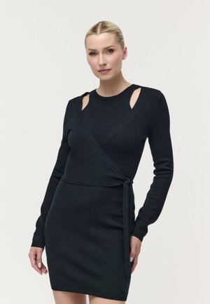 Vrouw met blond haar in een knot, die een zwarte jurk met lange mouwen, schouderuitsparingen en een ceintuur in de taille draagt.